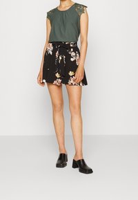 Grön ärmlös topp med spetsdetaljer, svart blommig kjol med knytmidja, och svarta klackskor med blockklack. Strukturerade tyger, blommönster.