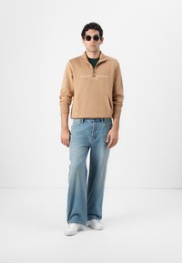 Hnedá mikina po zapínaní s logom, predná vrecková kapsa, voľné svetlomodré džínsy a biele tenisky; čierne slnečné okuliare dotvárajú outfit.