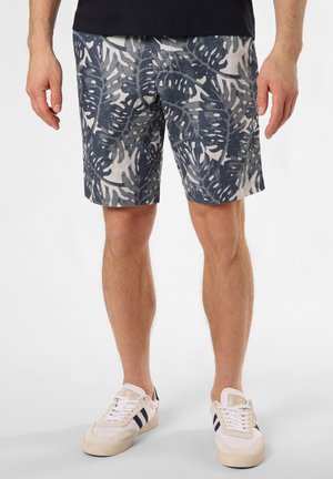 Mann in navy-blauen und weißen Shorts mit tropischem Blattmuster und weißen Sneakers mit schwarzen Streifen, der vor einem schlichten Hintergrund steht.