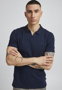 Marineblå poloskjorte laget av glatt bomullsstoff med klassisk krage, korte ermer og en subtil logo på brystet.