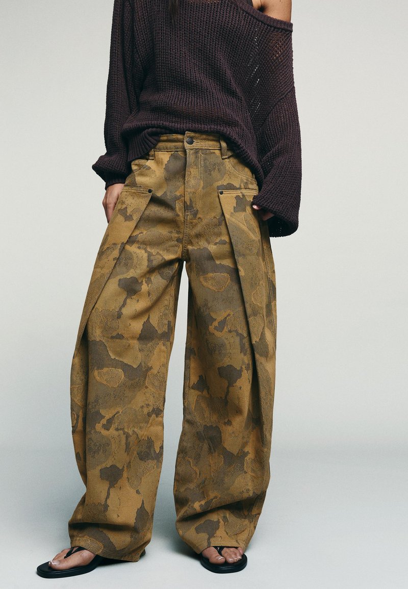 Personne portant un pantalon camouflage marron à taille haute et jambes larges, un pull en maille violet foncé oversize et des tongs noires sur un fond uni.