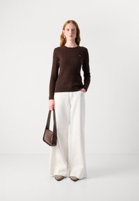 Pull en tricot brun à mailles, avec des manches longues, associé à un pantalon large crème. Le mannequin tient un petit sac à main brun, portant des chaussures aux tons neutres.