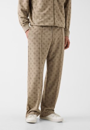 Homme portant un pantalon de survêtement beige à motifs et une veste zippée assortie, avec des baskets blanches, se tenant devant un fond uni.