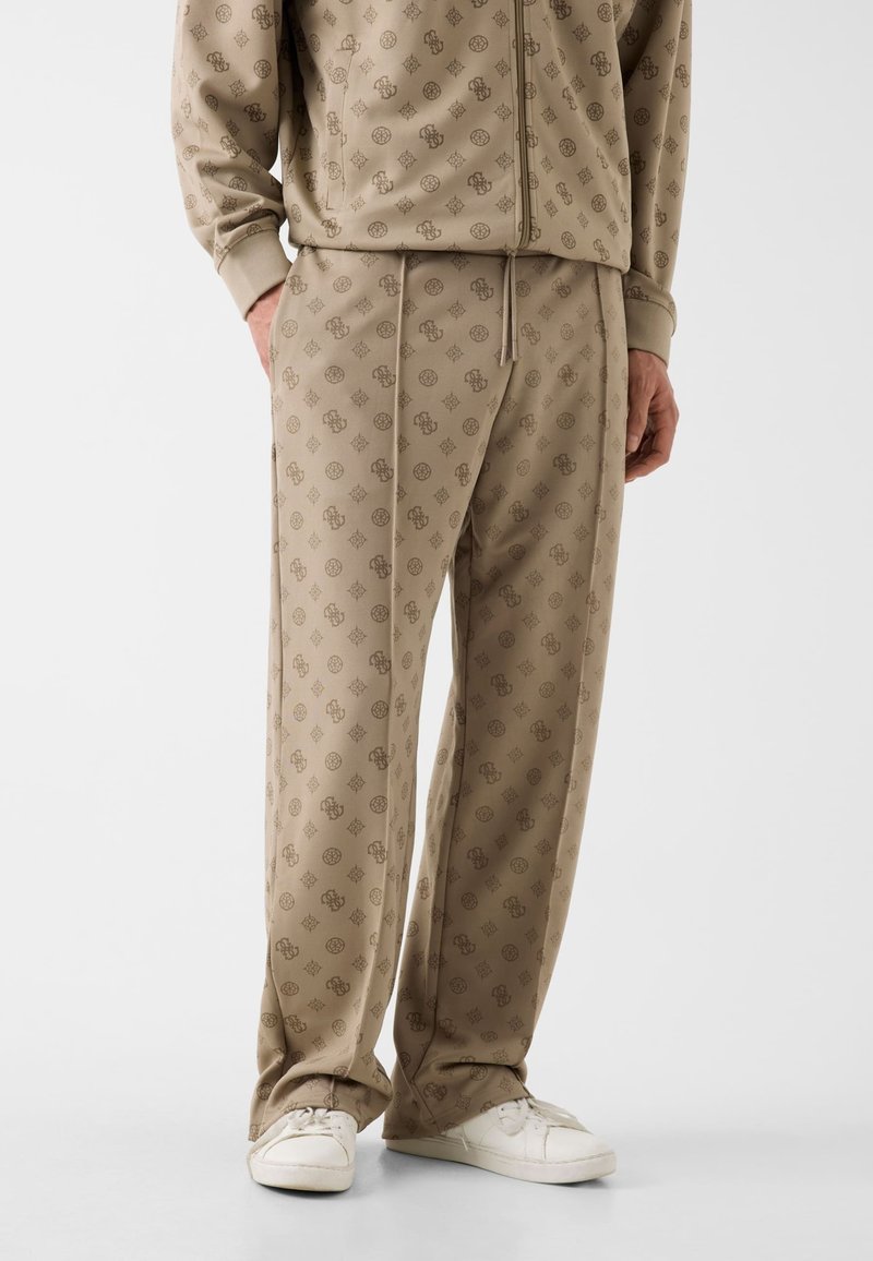 Mann trägt beige gemusterte Trainingshose und passende Reißverschlussjacke mit weißen Turnschuhen, steht vor einem einfarbigen Hintergrund.