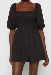 Musta lyhyt mekko kevyestä kankaasta, jossa on puffi hihat, neliönmuotoinen kaula-aukko ja kerätty elastinen vyötärö rentoa istuvuutta varten.