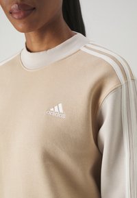 Béžová mikina s žebrovaným límcem, třemi bílými pruhy na ramenou a bílým vyšívaným logem Adidas na hrudi. Měkká textura látky.