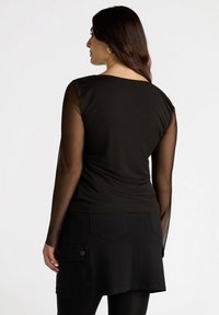 Top negro con mangas largas transparentes, que presenta un escote en forma de scoop. Combinado con una falda negra que tiene un bolsillo lateral y un detalle de botón.