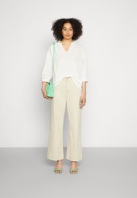 Blusa branca texturizada com decote em V, combinada com calças largas beiges. Segurando uma pequena bolsa de tiracolo verde-menta e usando sandálias verdes.