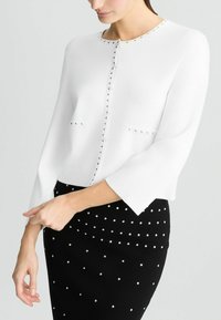 Cardigan bianco corto con dettagli in tono argento lungo il collo rotondo e la parte frontale, abbinato a una gonna a tubino nera decorata con gemme.