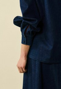 Mörk denimskjorta med lös passform, rynkad ärm och knappslutning, matchad med en kjol som framhäver en jämn textur.