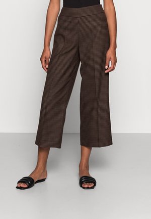 Pantalones anchos cortos marrones con cuadros, usados con sandalias negras tipo slide en una persona de pie contra un fondo liso.