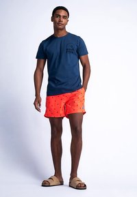 Tmavě modré bavlněné tričko s logem na hrudi, doplněné o jasně oranžové plavky s motivem malých teček a nášivkou.