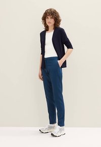 Tengerészkék kardigán fehér felső felett, türkiz nadrággal és szürke sportcipővel párosítva. A modell világos háttér előtt áll.