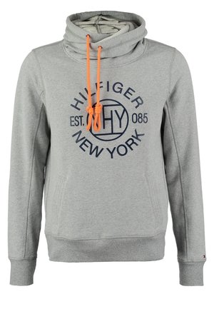 Mężczyzna w szarej bluzie z kapturem Tommy Hilfiger, czarnych podwiniętych spodniach, białych wsuwanych butach, trzymający brązową skórzaną torbę na sznurkach.