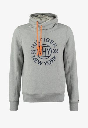 Mann trägt grauen Tommy Hilfiger Hoodie, schwarze hochgekrempelte Hosen, weiße Slip-On-Schuhe und hält eine braune Ledersacktasche mit Kordelzug.