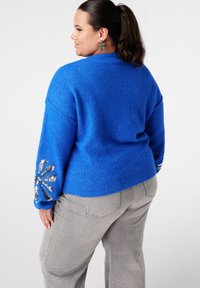 Femme posant de profil, portant un pull bleu vif avec des motifs de fleurs en sequins argentés sur les manches et un jean gris clair.