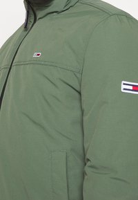 Grüne gesteppte Jacke mit schwarzem Reißverschluss, mit einer Brusttasche und einem Logoaufnäher auf dem Ärmel mit roten, weißen und blauen Akzenten.