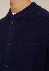 Cardigan en tricot bleu marine présentant un motif texturé, avec un col côtelé et quatre boutons noirs texturés. Vue en gros plan du tissu.
