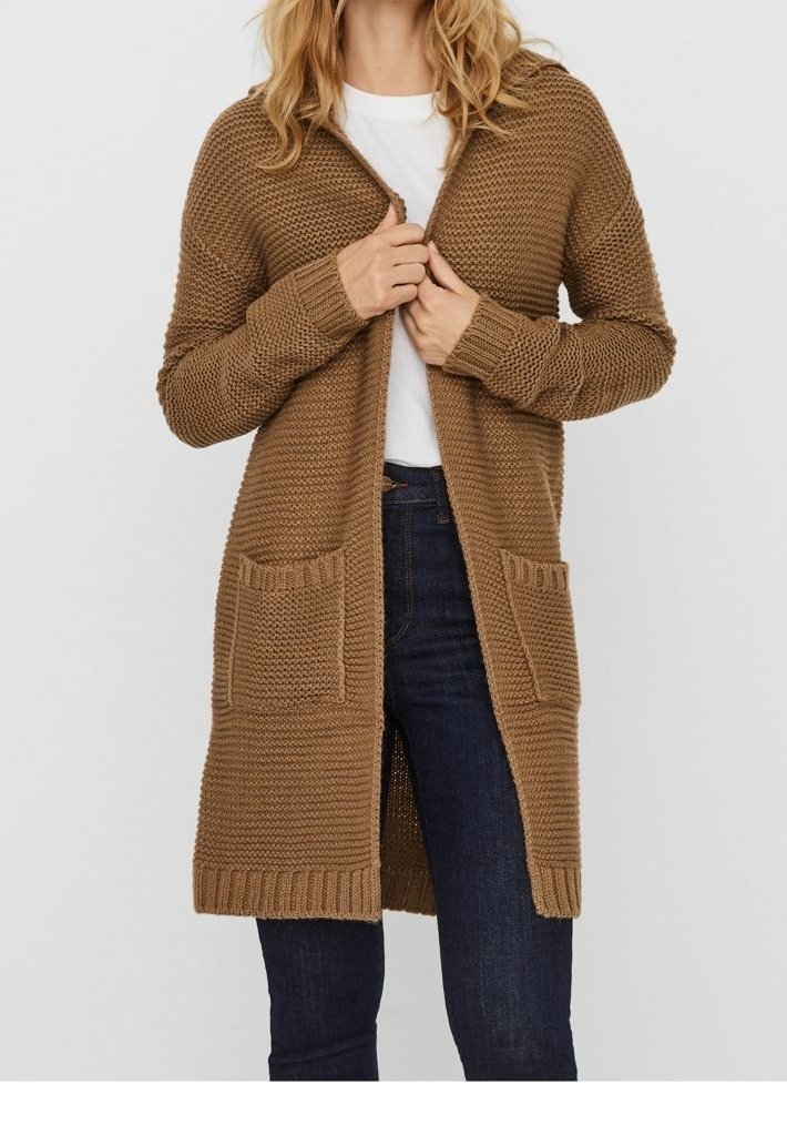 Femme portant un long cardigan marron en tricot sur une chemise blanche et un jean bleu foncé, tenant le cardigan fermé au niveau de la poitrine.