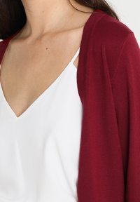 Burgundy cardigan made of soft knit fabric, open front design, worn over a white v-neck top, highlighting smooth texture and relaxed fit.