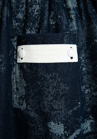 Tejido de mezclilla con un patrón texturizado en azul oscuro, con una etiqueta blanca con texto en relieve y cierre a presión.