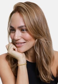 Złota bransoletka z wzorem z koralików na nadgarstku; modelka ma długie, proste, jasnobrązowe włosy, nosi czarny top bez rękawów i delikatnie się uśmiecha.