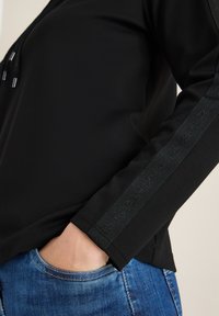 Czarny longsleeve z dekoltem na sznurki i teksturowymi akcentami wzdłuż rękawów. Połączony z niebieskimi dżinsami.