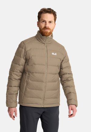 Mann mit rotem Bart, der eine hellbraune Steppjacke mit hohem Kragen trägt und vor einem einfarbigen Hintergrund steht.