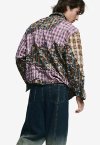 Kötött kabát lila, barna és kék kockás minták keverékével. Jellegzetes hullám mintázattal és bő szabású sötét farmernadrággal.