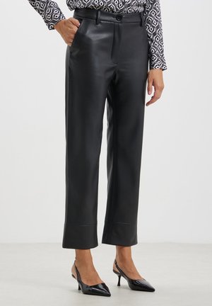 Femme portant un pantalon en cuir noir droit, une blouse à manches longues à motifs, et des escarpins slingback noirs à bouts pointus, debout sur un sol blanc.