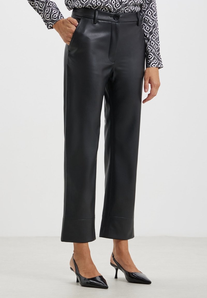 Femme portant un pantalon en cuir noir droit, une blouse à manches longues à motifs, et des escarpins slingback noirs à bouts pointus, debout sur un sol blanc.