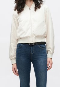 Veste bomber beige clair avec zip à l'avant, poignets et ourlet côtelés. Portée avec un jean bleu foncé, coupe slim et lavage moyen.