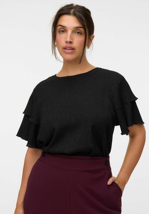 Femme aux cheveux foncés portant un haut noir à manches courtes avec des volants et un pantalon taille haute bordeaux, la main dans la poche, sur un fond clair uni.