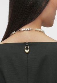 Μαύρο ένδυμα με ευρύ λαιμόκοψη, με φερμουάρ στο πίσω μέρος, διακοσμημένο με χρυσή λεπτομέρεια και αλυσίδα με μαργαρίτα που φέρει την επιγραφή "HUGO".