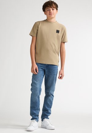 T-shirt en coton beige avec col rond et patch logo noir, associé à un jean bleu coupe slim et des baskets blanches. Design simple et coupe décontractée.