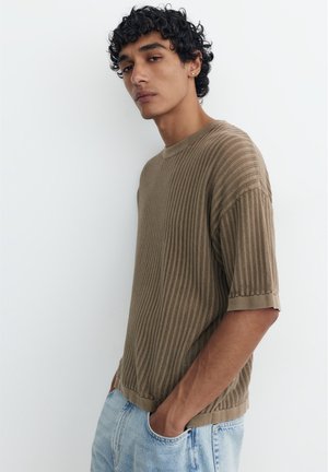 Junger Mann mit lockigem dunklem Haar, der ein strukturiertes kurzärmeliges beiges Strickshirt und hellblaue Jeans trägt, steht vor einem weißen Hintergrund.