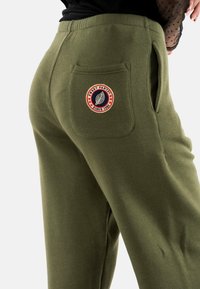 Pantalones de chándal verde oliva con un tejido texturizado, cintura elástica, bolsillos laterales y un parche redondo con un diseño de hoja y texto.