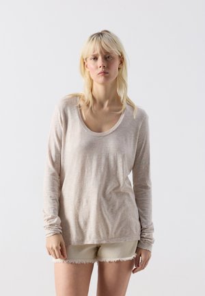 Mladá žena se světlými vlasy, oblečená v béžovém triku s dlouhým rukávem a světle zbarvených roztřepených šortkách, stojící před jednobarevným bílým pozadím.