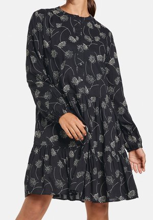 Robe noire à manches longues avec un motif floral blanc, boutons sur le devant, et une jupe à volants froncée, portée par une personne debout.