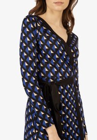 Blau-schwarzes Wickelkleid mit dreieckigen Mustern in Beige und Blau, langen Ärmeln, einem tiefen V-Ausschnitt und einem schwarzen Gürtel in der Taille.