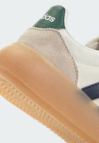 Zapato de piel blanca con tacón texturizado verde, detalles en ante beige y suela de goma clara con un patrón en zigzag. Detalle de raya azul incluido.