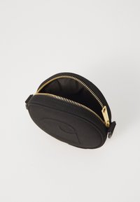 Black circular zipper pouch made of fabric, featuring a gold zipper and a textured face with a subtle logo. Interior is lined and dark.