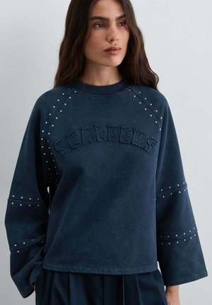 Mujer con el pelo largo y oscuro que lleva una sudadera azul marino con mangas anchas y "SCALPERS" bordado en el pecho.