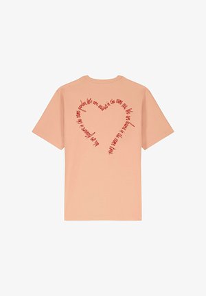 T-shirt en coton de couleur pêche avec un col rond. Le dos présente un texte rouge disposé en forme de cœur, exprimant un message inclusif.