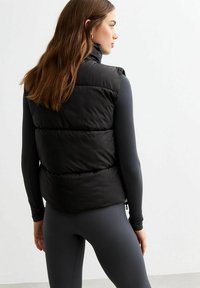 Chaleco acolchado negro con cuello alto, que presenta compartimentos y una textura suave, usado sobre un atuendo oscuro ajustado. Diseño simple y funcional.