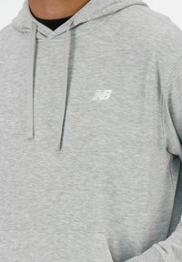 Helle graue Kapuzenjacke aus weichem Stoff, mit einer Vordertasche, Kordeln und einem kleinen weißen Logo auf der Brust.
