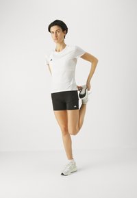 Weißes Sportshirt mit drei schwarzen Streifen, schwarze Shorts und hellfarbene Turnschuhe. Die Person dehnt das Bein und zeigt damit das Design und die Passform.