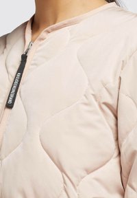 Light pink quilted jacket with a zip closure, featuring rounded seams and a contrasting dark label on the zipper. Soft, smooth texture.