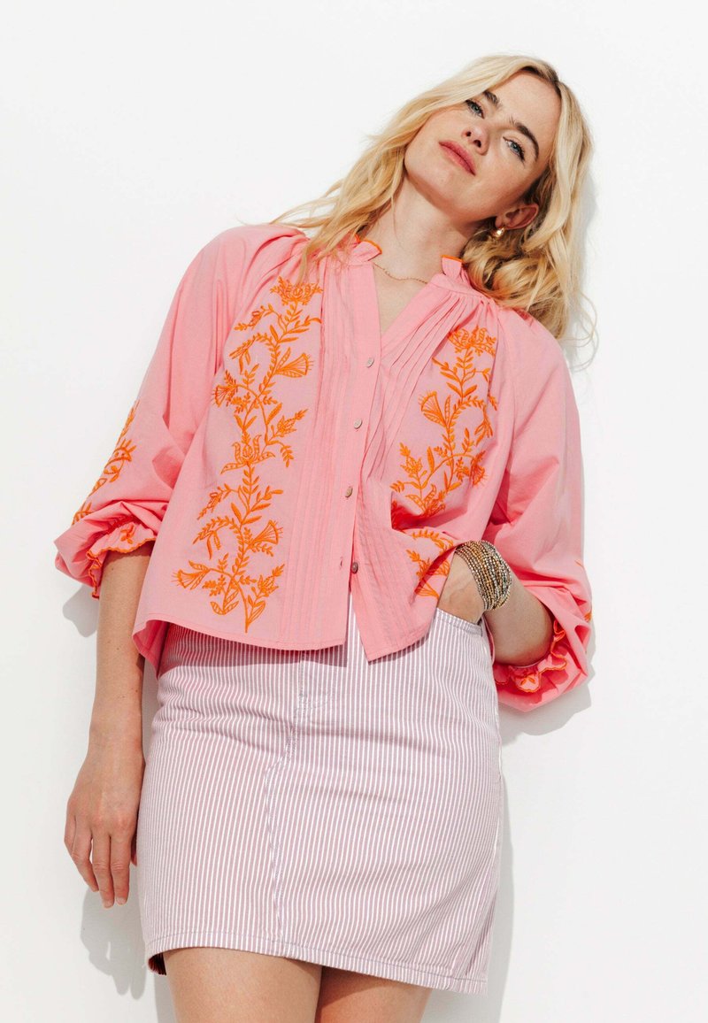 Blouse rose à manches courtes avec broderie florale orange, détails en boutons et devant plissé. Portée avec une mini-jupe rayée.