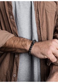 Juodas lavos akmens apyrankė su apvaliais karoliukais, papuošta sidabro akcentu. Nešiojama ant riešo, derinama su pilku marškinėliu ir rudais veliūro marškiniais.